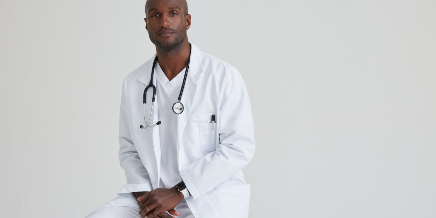Black male healthcare professional wearing a white lab coat and stethoscope, seated against a plain light grey background.