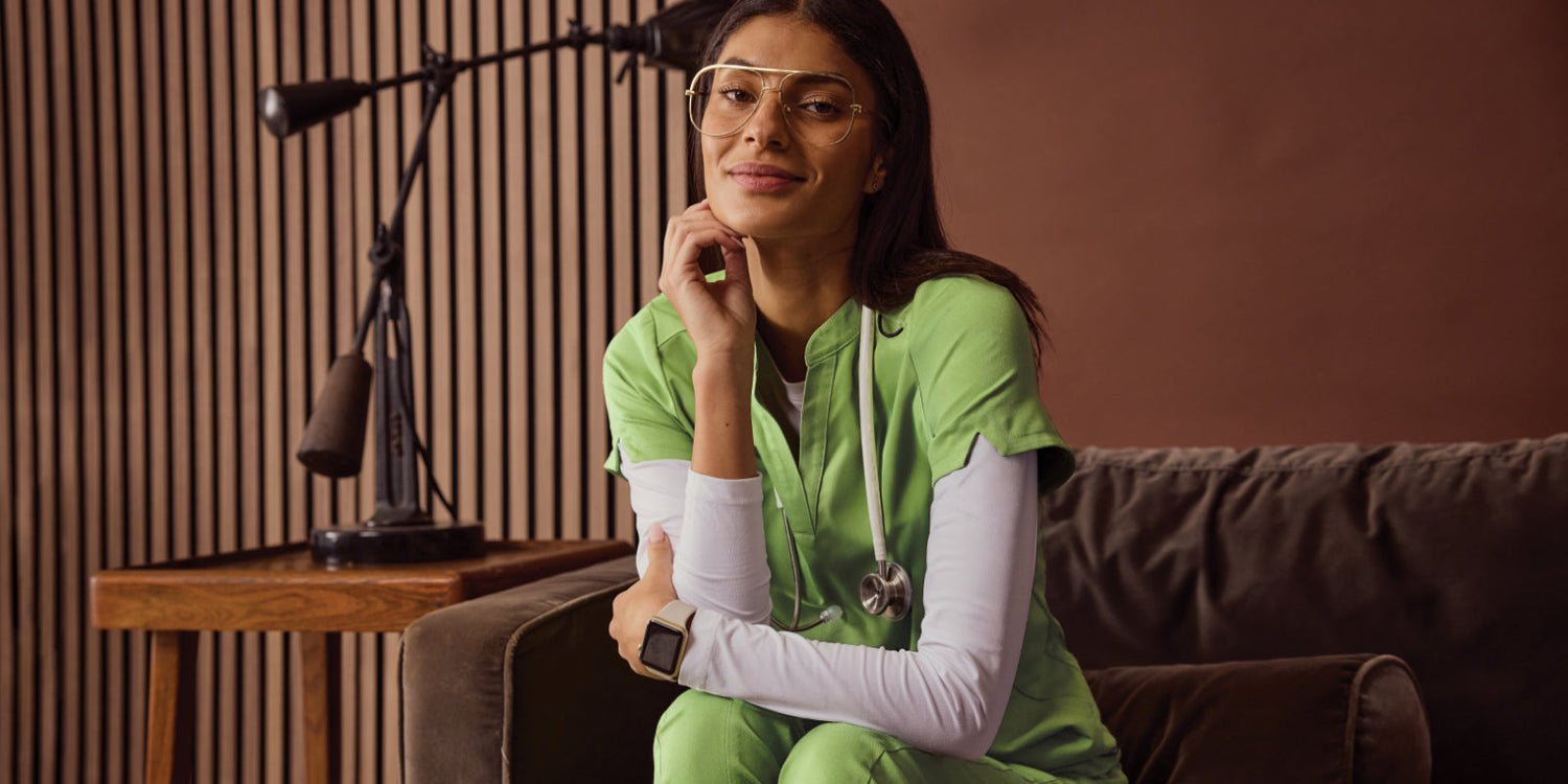 Woman sitting on a couch wearing lime green scrub pants and top layered over a white long-sleeve underscrub shirt, smiling in a warmly lit room.