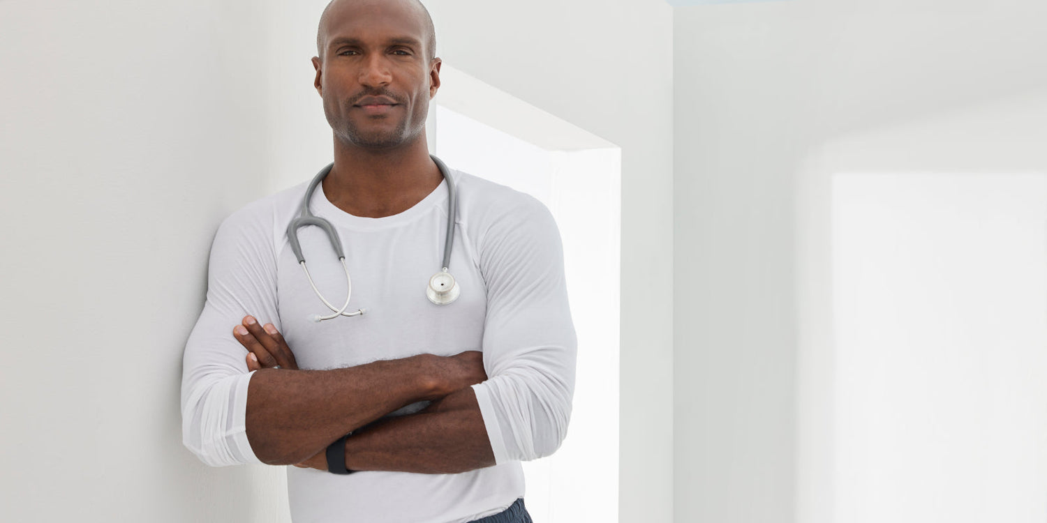 Male medical professional wearing a fitted long sleeve white underscrub shirt with a stethoscope, standing confidently in a bright, modern clinical setting.
