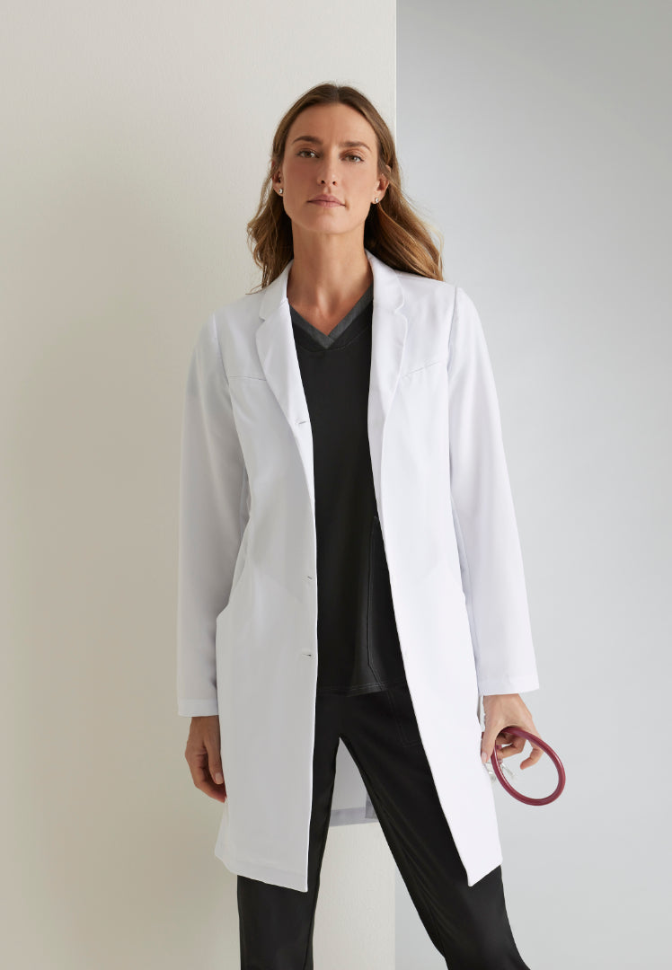 Woman wearing a white lab coat over black scrubs, holding a stethoscope, standing against a neutral background.