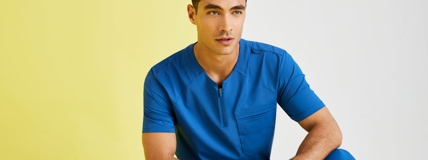 Male model wearing a bright blue scrub top, seated in front of a yellow and white background.
