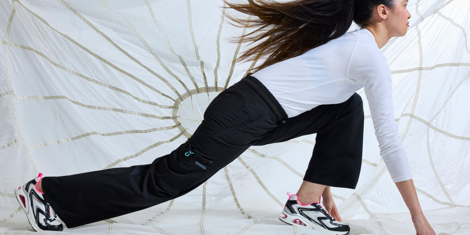 Woman stretching in black medical scrub pants and sneakers, wearing a white long-sleeve top, in front of a white parachute backdrop.