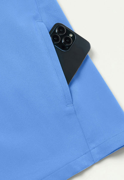 Detiled view of a Jaanuu Platt scrub top in ciel blue with a black smartphone peeking out of a packet against a white background