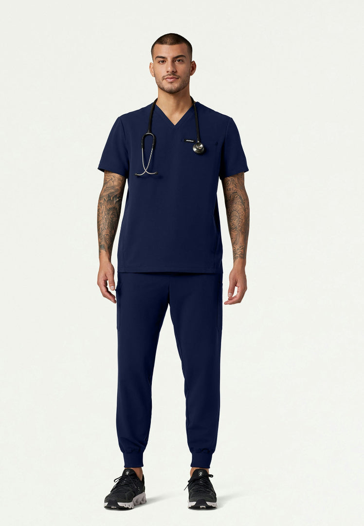 Full body view of a man wearing a navy blue Jaanuu medical scrub set and a stethoscope on a white background