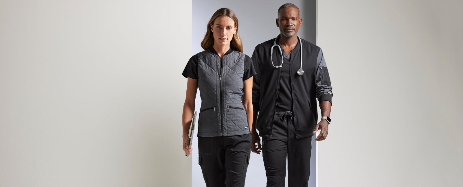 Two healthcare professionals, a woman on the left and a man on the right, confidently model the latest in medical scrubs from 'The Uniform Store'. The woman is wearing a fitted, quilted vest over her scrubs, while the man dons a sleek, black zippered jacket atop his. They both carry stethoscopes, symbolizing their readiness to serve. Text overlay reads 'Healthcare Apparel Experts - Join the community of healthcare heroes who trust our scrubs and coats.