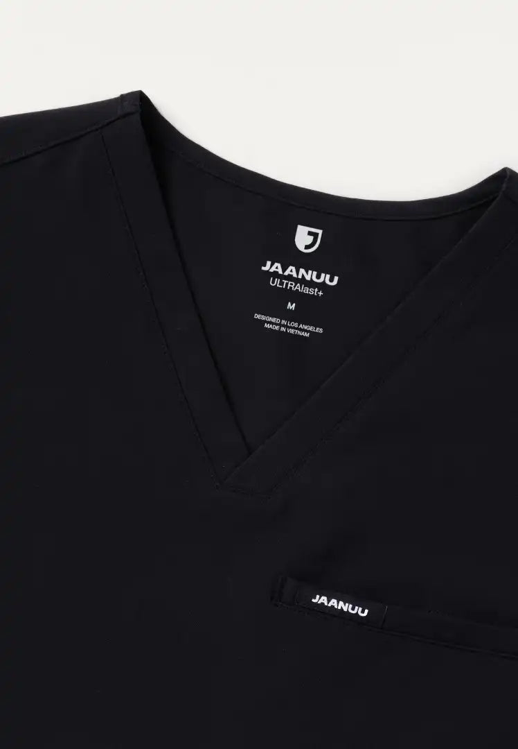 Detailed view of a Jaanuu men's Platt V-neck scrub shirt in black with Jaanuu branding on a white background
