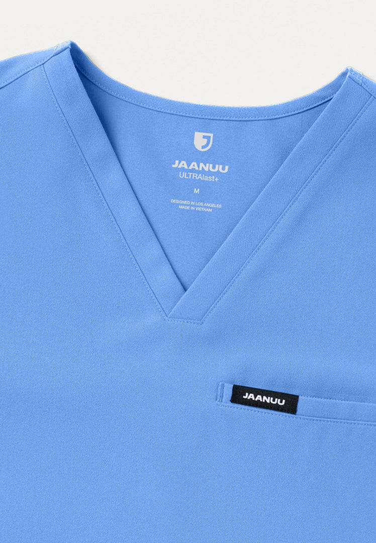 Detailed view of a men's Jaanuu Platt v-neck scrub top in ciel blue with Jaanuu branding on a light gray background