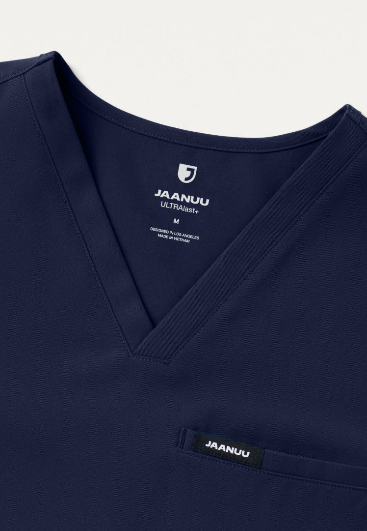 Detailed view of a navy blue Jaanuu Platt scrub top with 'Jaanuu' branding on a light gray background