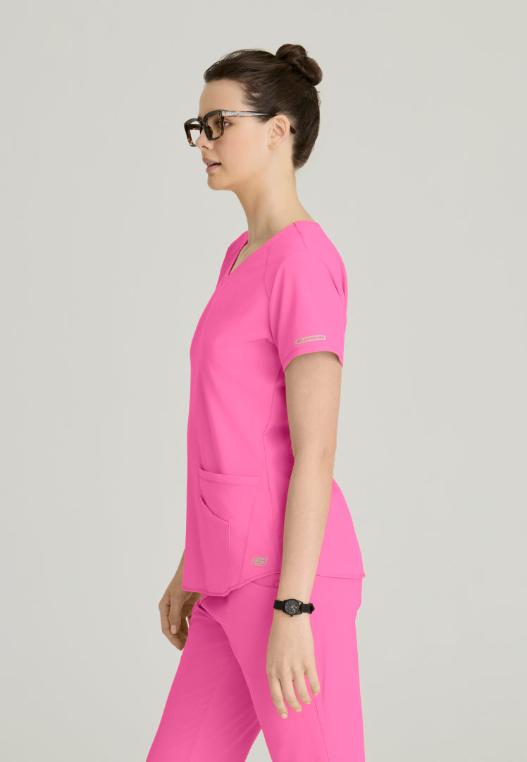 Side view of a woman wearing a Skechers SK101 Passion Pink scrub shirt on a light gray background