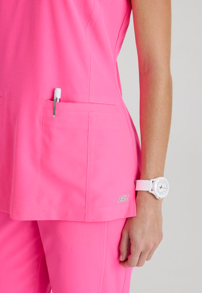 Pocket detail view of a woman wearing a Skechers SKT259 Passion Pink scrub shirt with a white watch on a light gray background