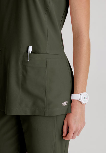 Pocket detail view of a woman wearing a Skechers olive green SKT259 scrub shirt with a white watch on a light gray background