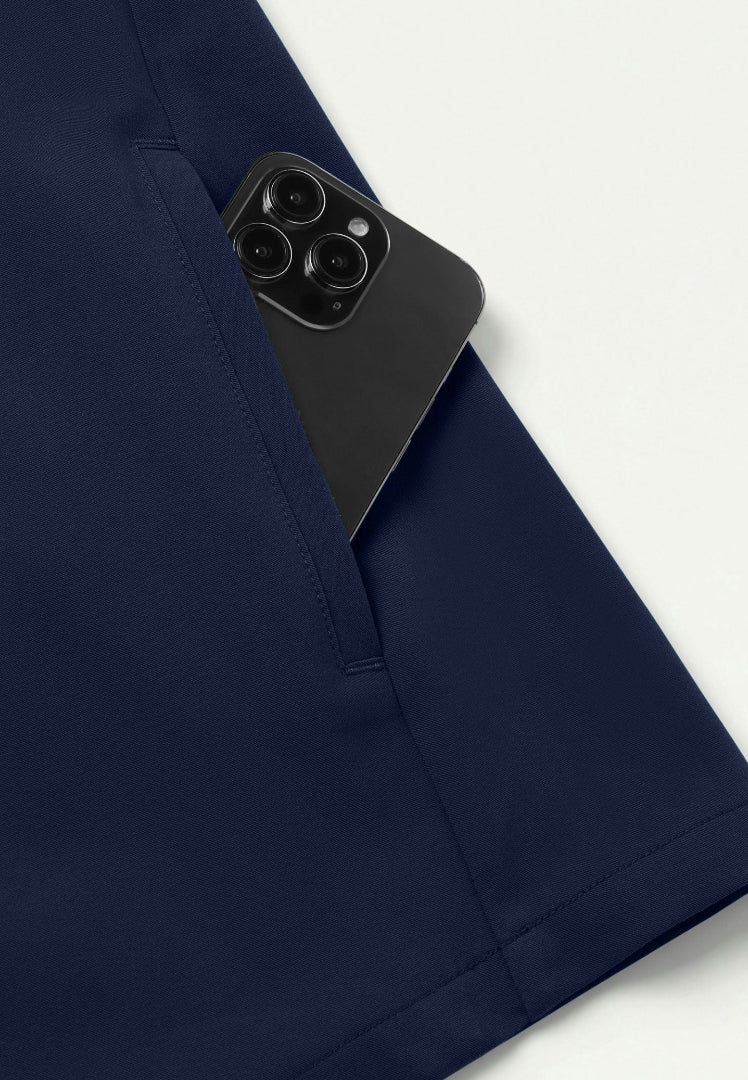 Detiled view showing a Jaanuu navy blue scrub top with a black smartphone peeking out of it's pocket on a white background